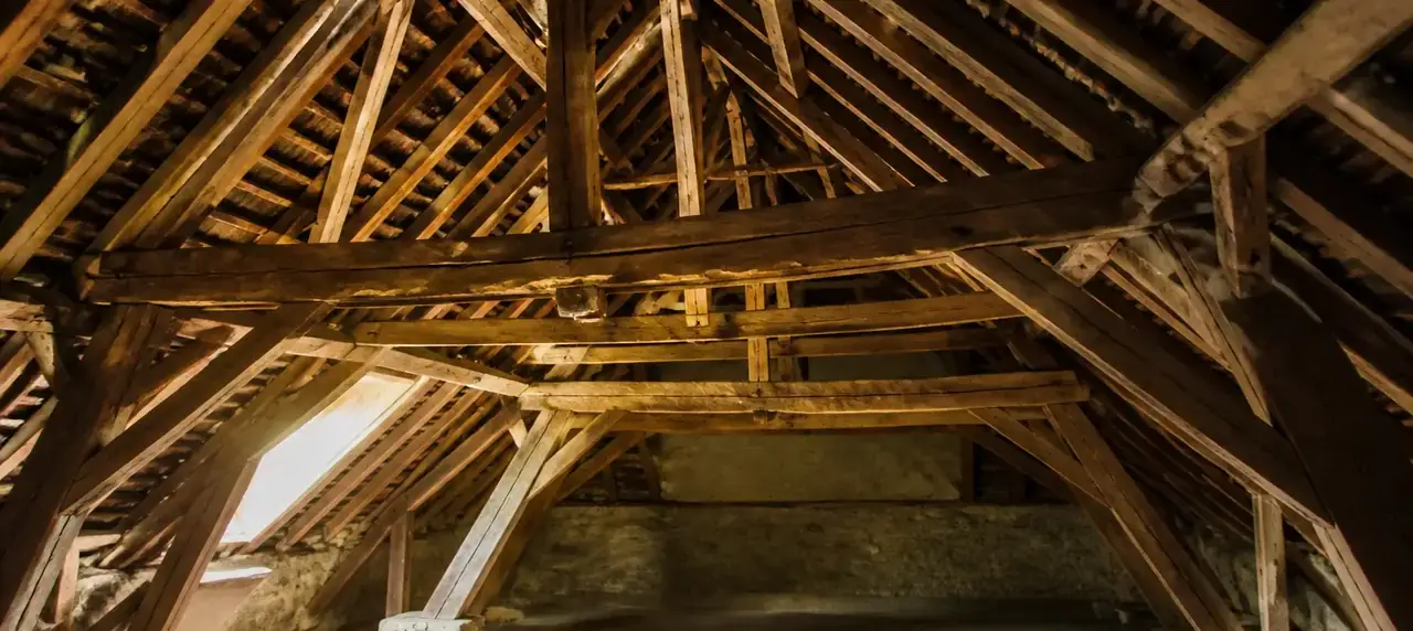 Charpente traditionnelle en bois vue de l’intérieur des combles, avec ses fermes, ses pannes et ses chevrons éclairés par une lucarne