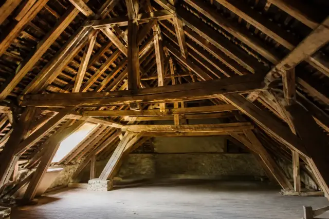 Charpente traditionnelle en bois vue de l’intérieur des combles, avec ses fermes, ses pannes et ses chevrons éclairés par une lucarne