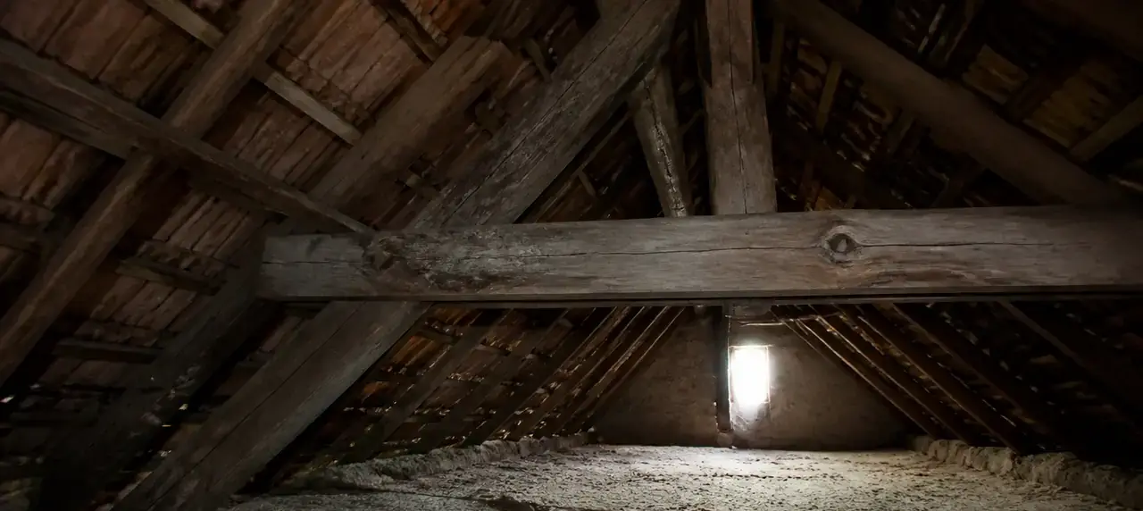 Combles d’une maison ancienne avec chevrons et pannes en bois, lampe de chantier posée sur un plancher