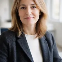 Portrait de Clémence Aubriot, cheveux châtain clair mi-longs, veste de laine gris ardoise sur un pull écru, sourire discret, fond de pièce claire et floue