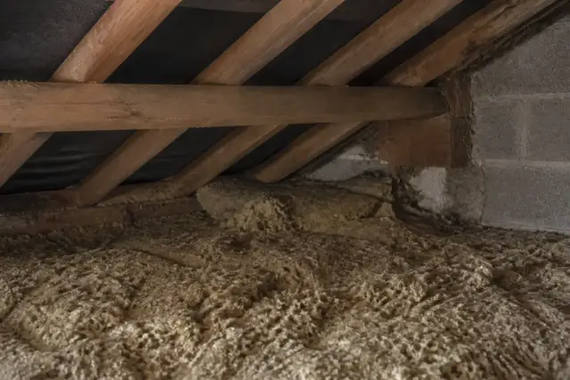 Combles perdus avec laine minérale arrachée et tassée près d’un chevron, lumière d’une lampe torche