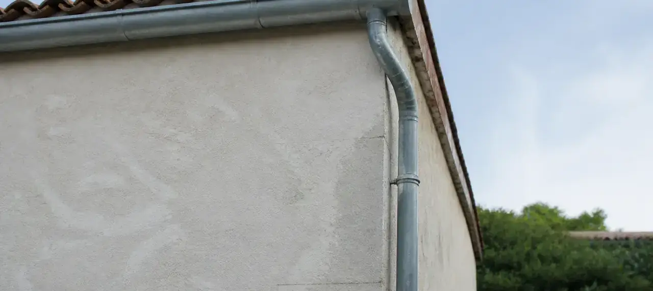 Angle de toit en tuiles avec une gouttière en zinc et sa descente, vu du sol contre un mur enduit clair
