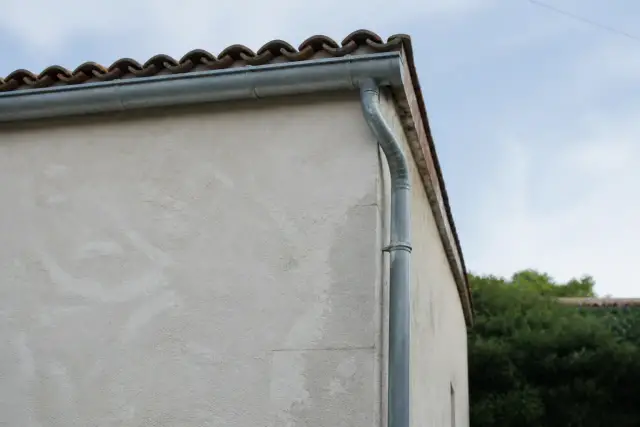 Angle de toit en tuiles avec une gouttière en zinc et sa descente, vu du sol contre un mur enduit clair