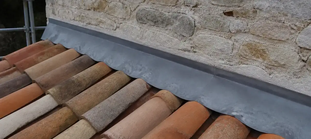 Bande de solin en zinc engravée dans un mur de pierre, au-dessus d’un rang de tuiles en terre cuite