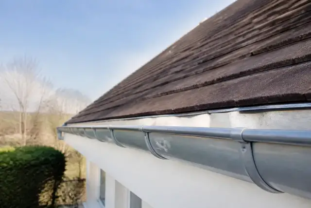 Toit de tuiles plates propres sous un ciel clair, avec une gouttière en zinc nettoyée au premier plan