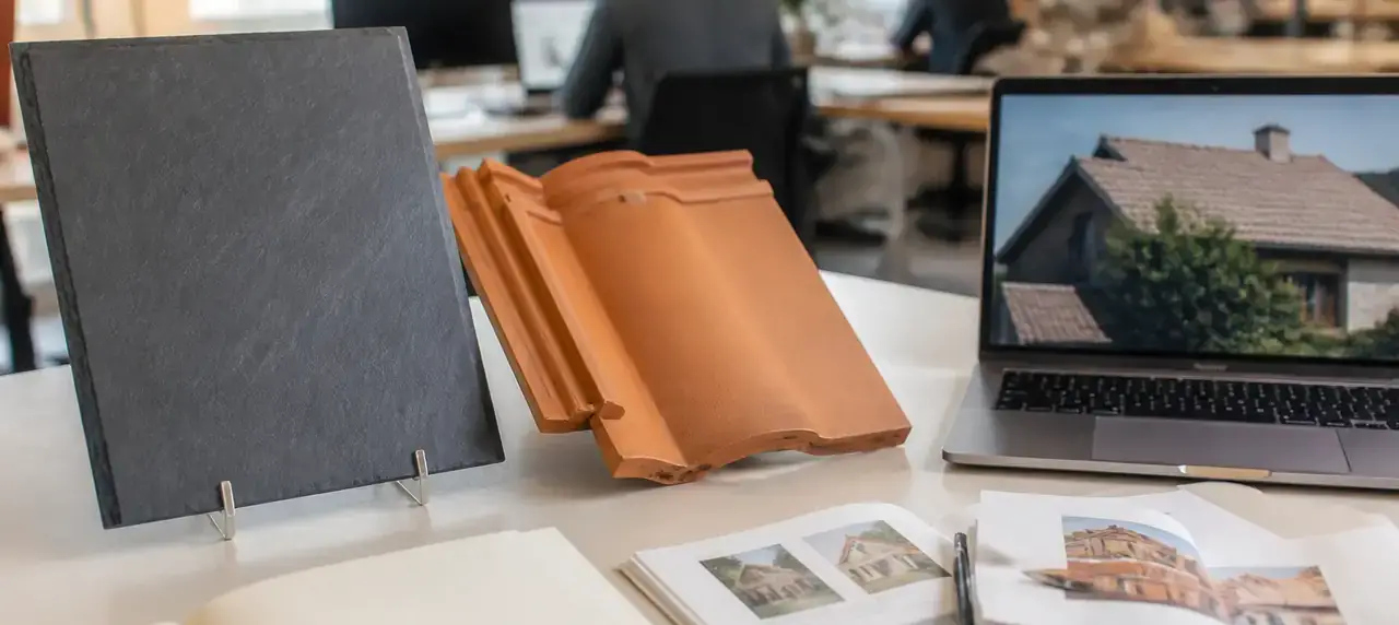 Bureau de rédaction clair avec des carnets ouverts, un échantillon d’ardoise et une tuile posés à côté d’un ordinateur
