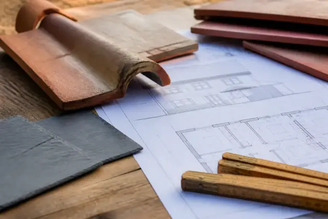 Plans de maison posés sur une table de bois avec des échantillons de tuile et d’ardoise et un mètre pliant