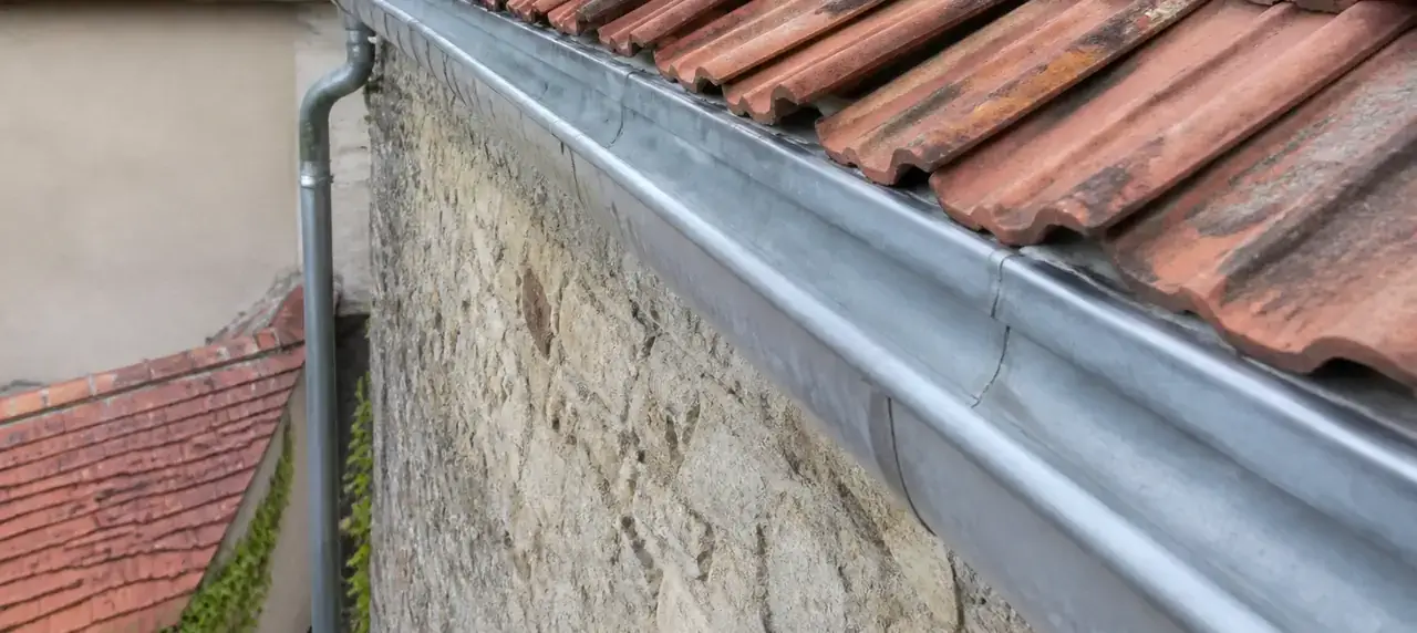 Chéneau en zinc posé en tête d’un mur de pierre séparant deux jardins, descente d’eau côté maison